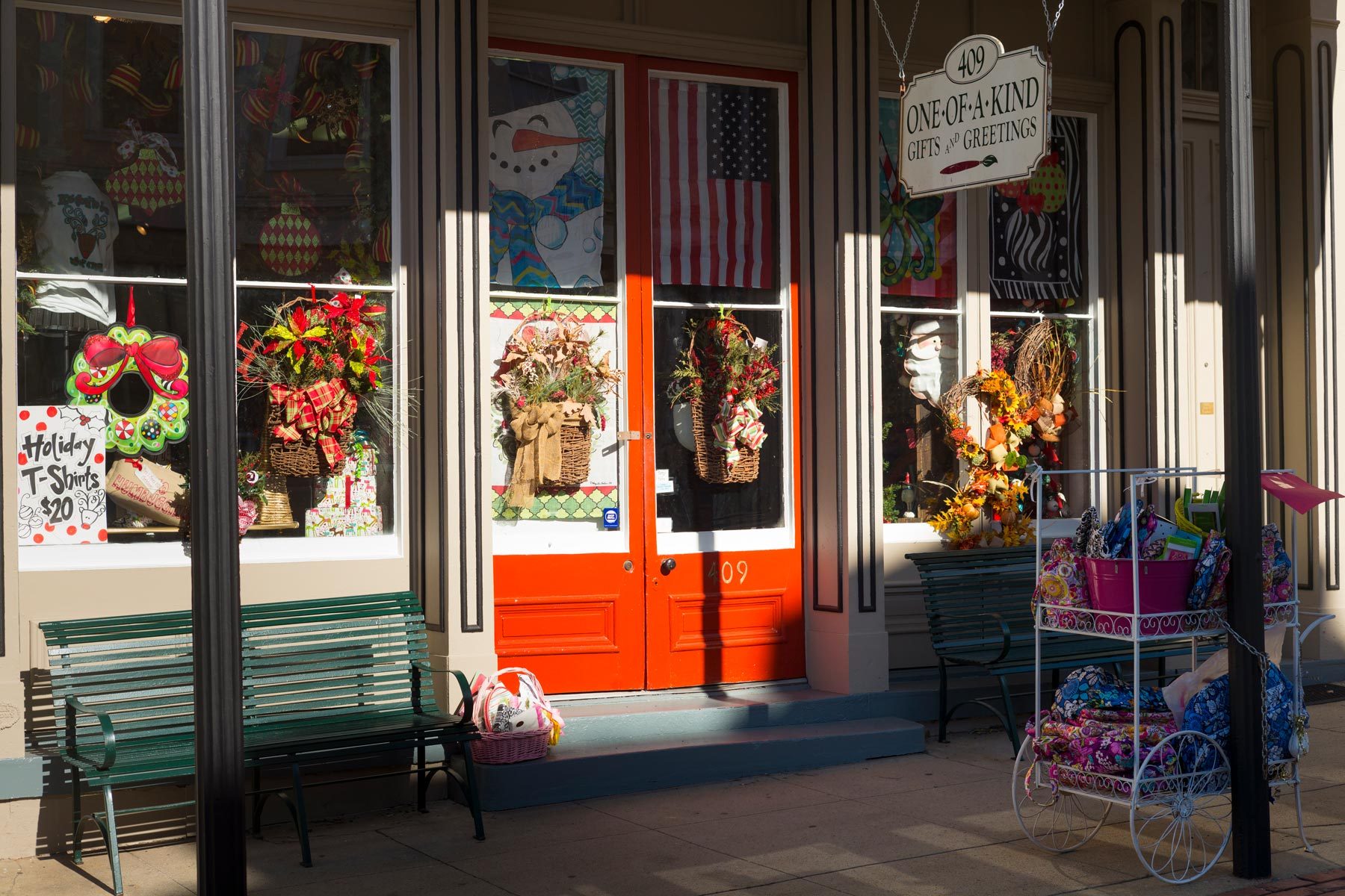 Storefront displays festive wreaths and decorations; "Holiday T-Shirts $20." Sign reads "409 One-of-a-Kind Gifts and Greetings," amidst benches and merchandise cart.