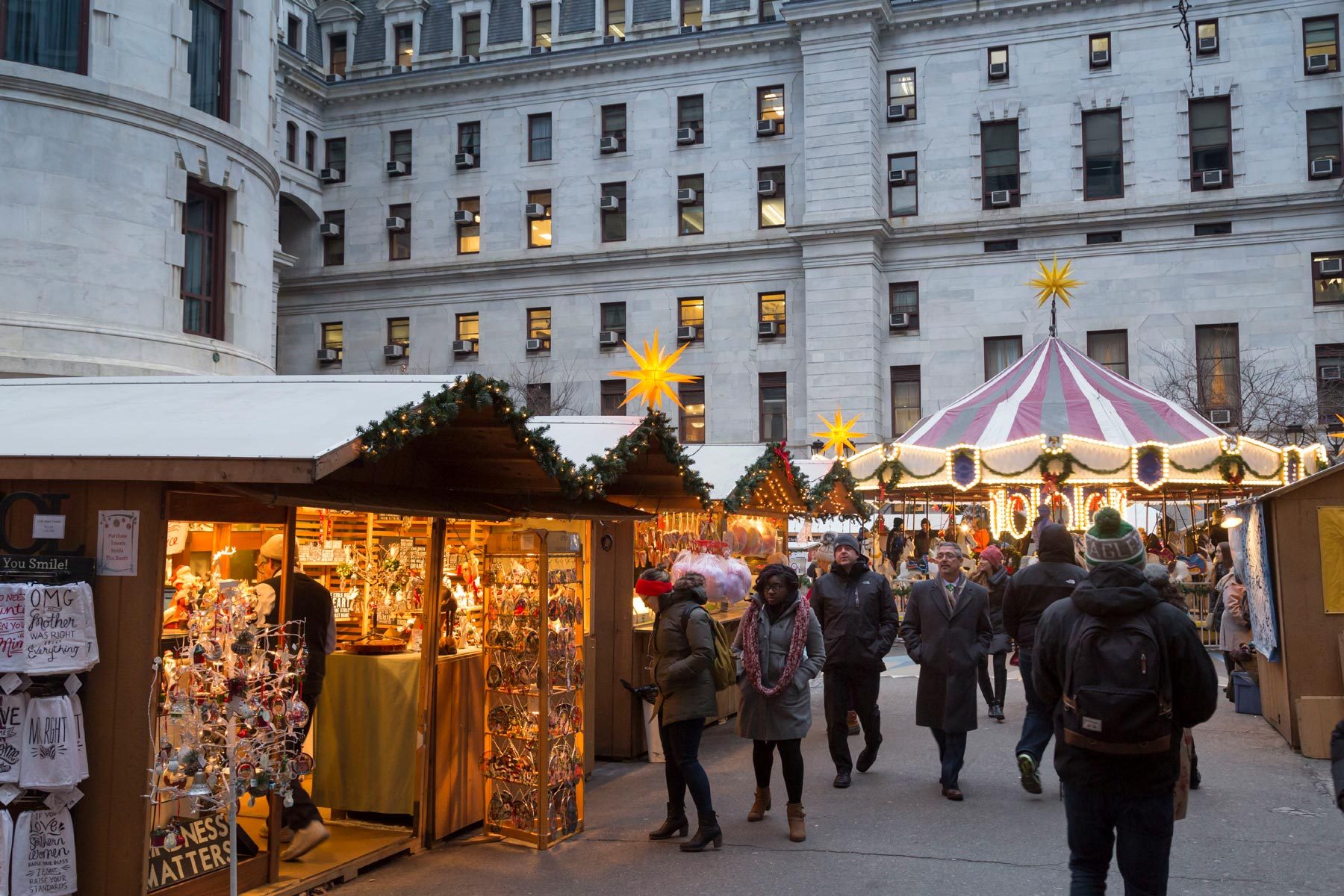 Market stalls display ornaments; people walk past. A festive carousel spins nearby, surrounded by decorative lights and a grand, historic building.