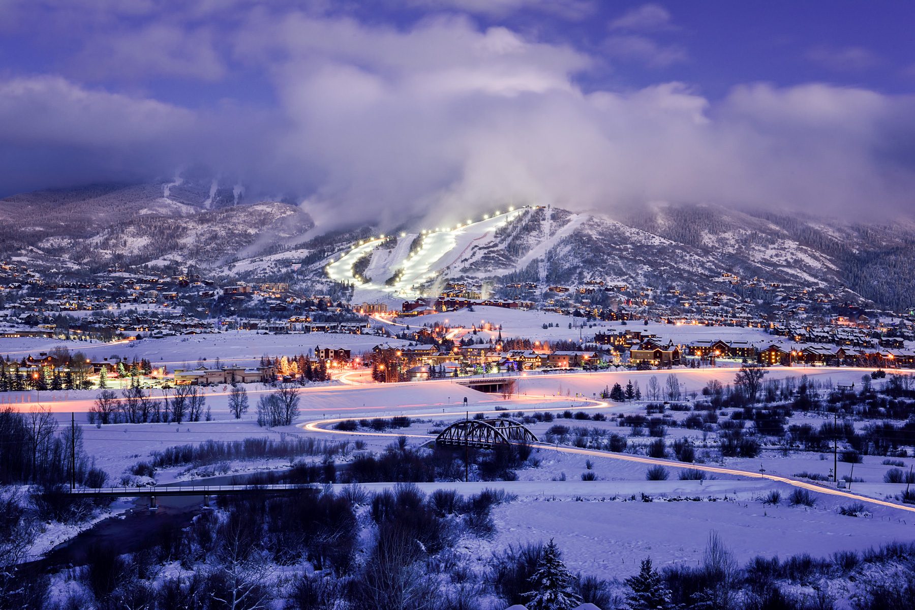 Snow-covered town at dusk, lights illuminate ski slopes and buildings, nestled within mountains under a cloudy sky.