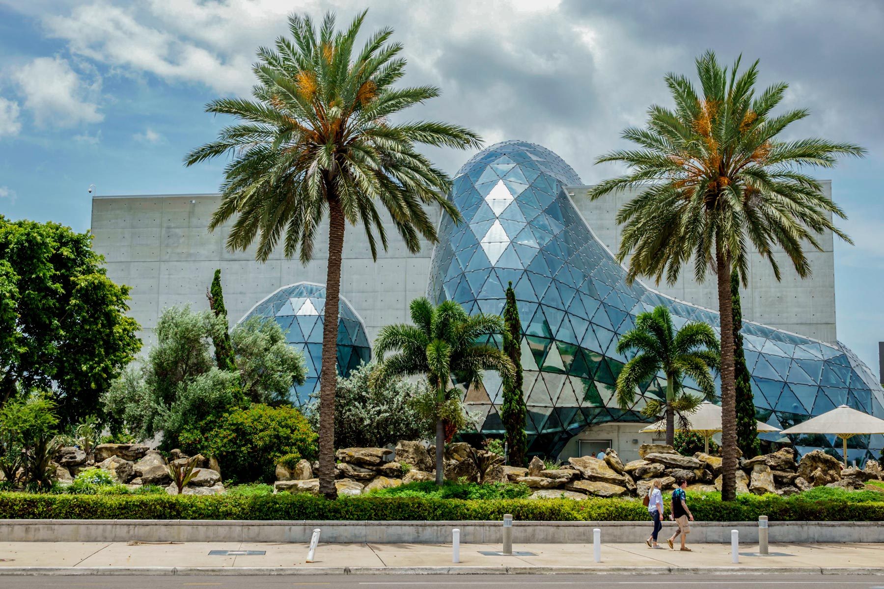Glass geodesic dome reflects sunlight, surrounded by palm trees and greenery. Two people walk on the sidewalk in front.