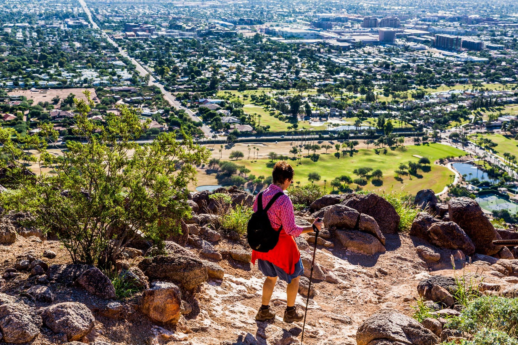 A hiker descends a rocky trail overlooking a sprawling cityscape with green golf courses and urban sprawl in the distance.