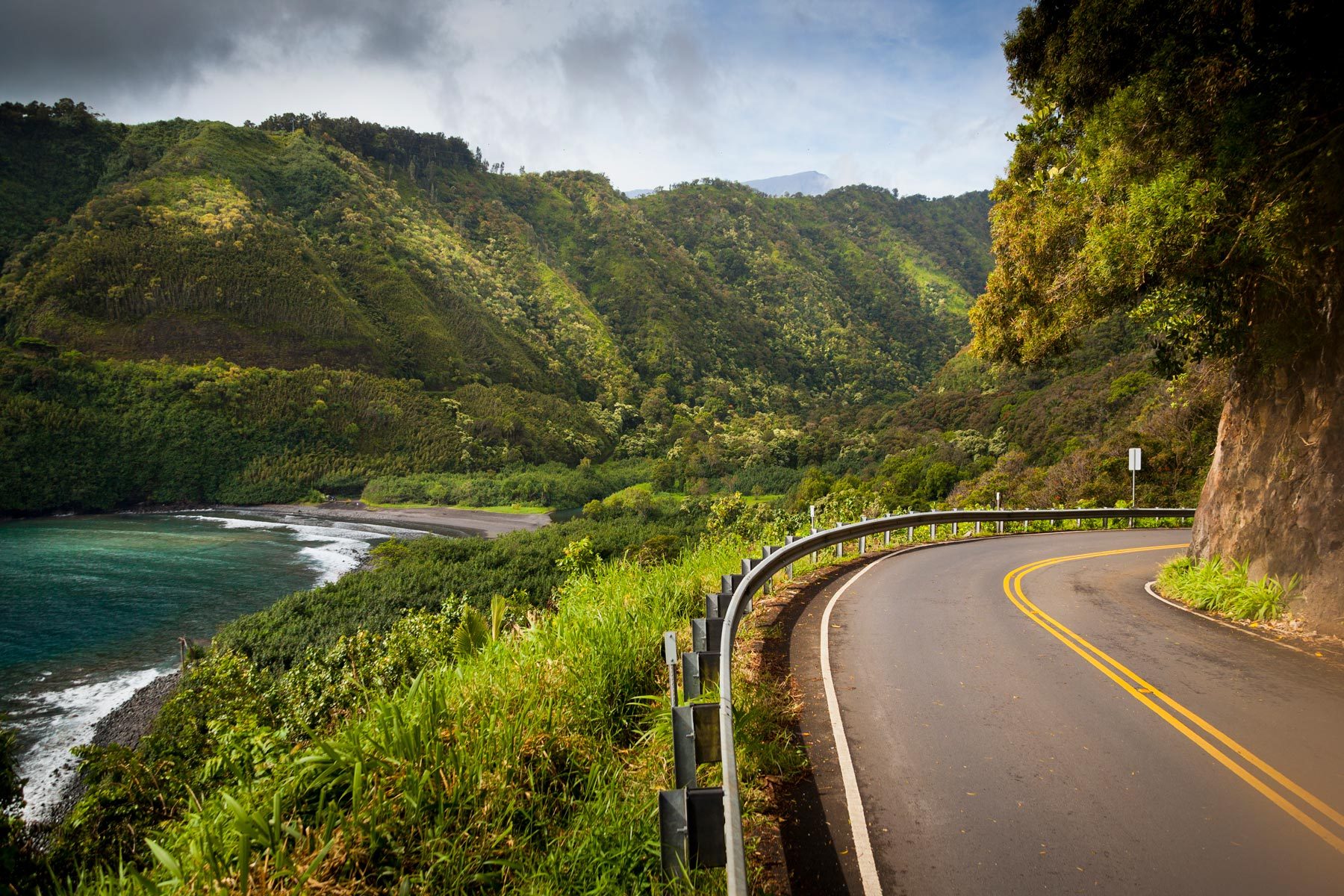 A winding road curves beside lush, green mountains and a tranquil bay under a cloudy sky.