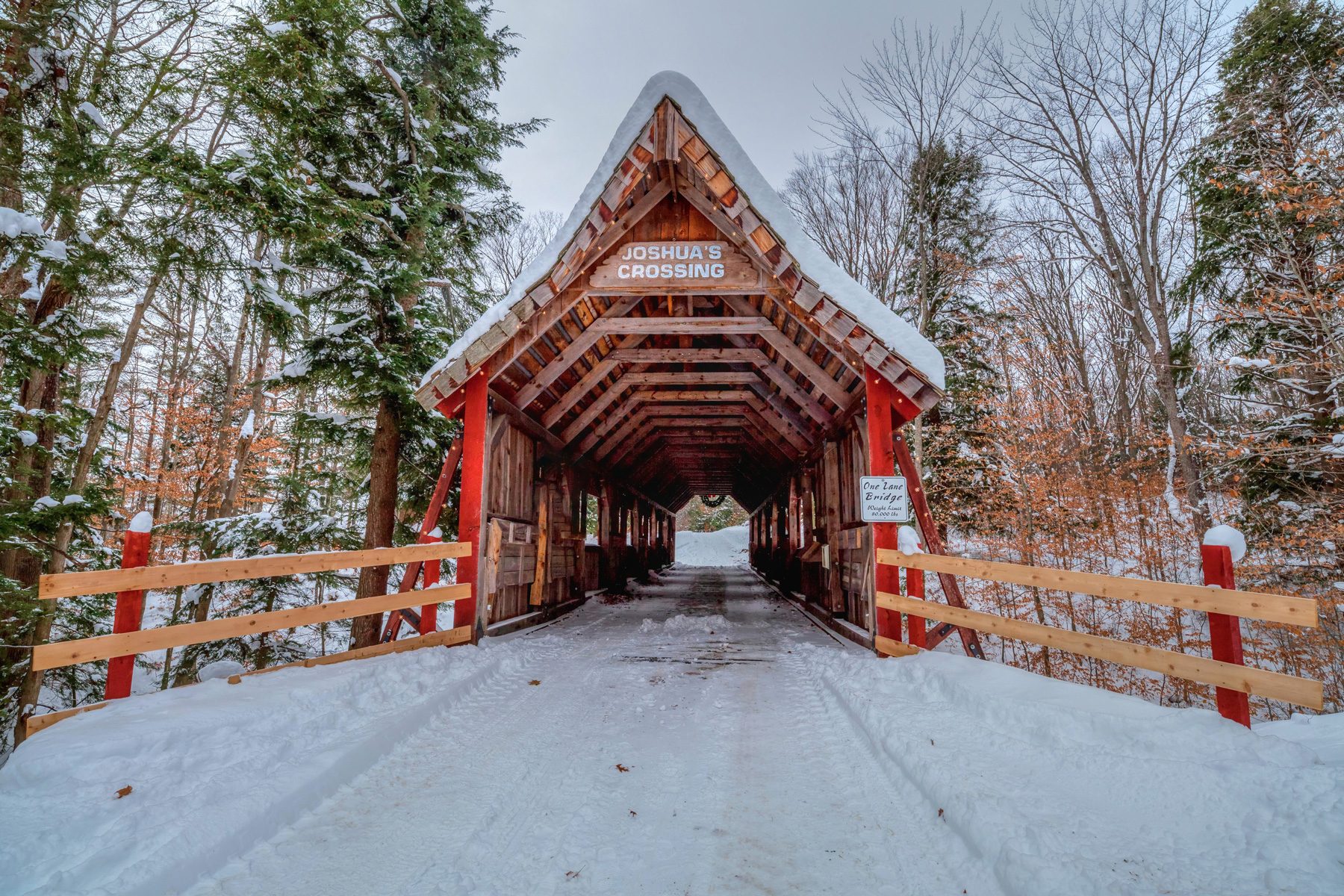 Snow-covered wooden bridge stands in snowy forest, entrance marked "JOSHUA'S CROSSING" and sign says "One Lane Bridge." Trees line the path.