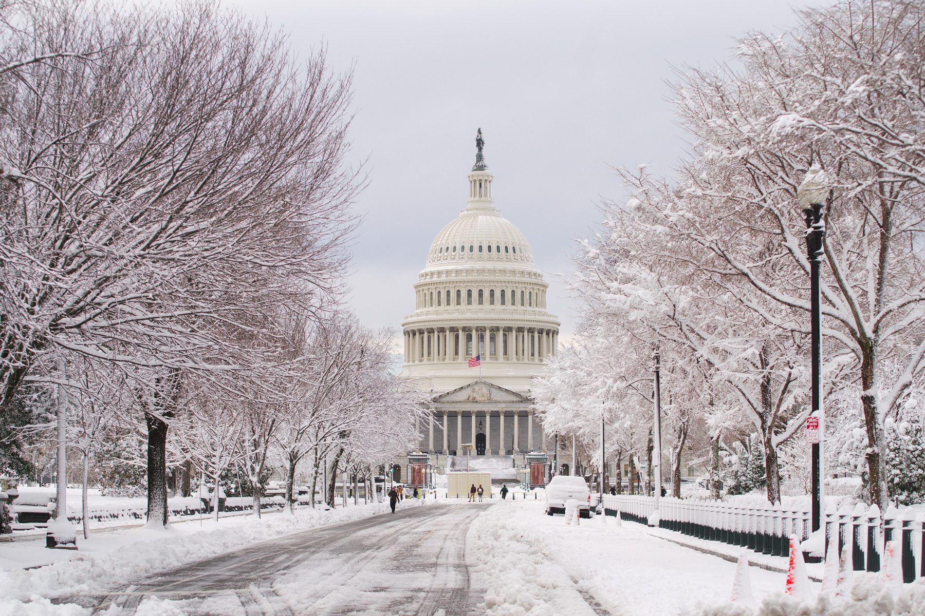 The Capitol building stands amid snow-covered trees and paths; an American flag flies in a wintry landscape.