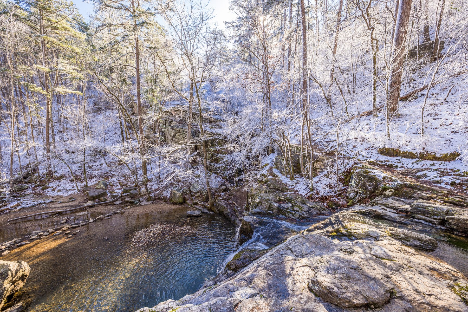Waterfall gently cascades over rocks, surrounded by frosty trees in a snow-dusted forest, under a clear blue sky.