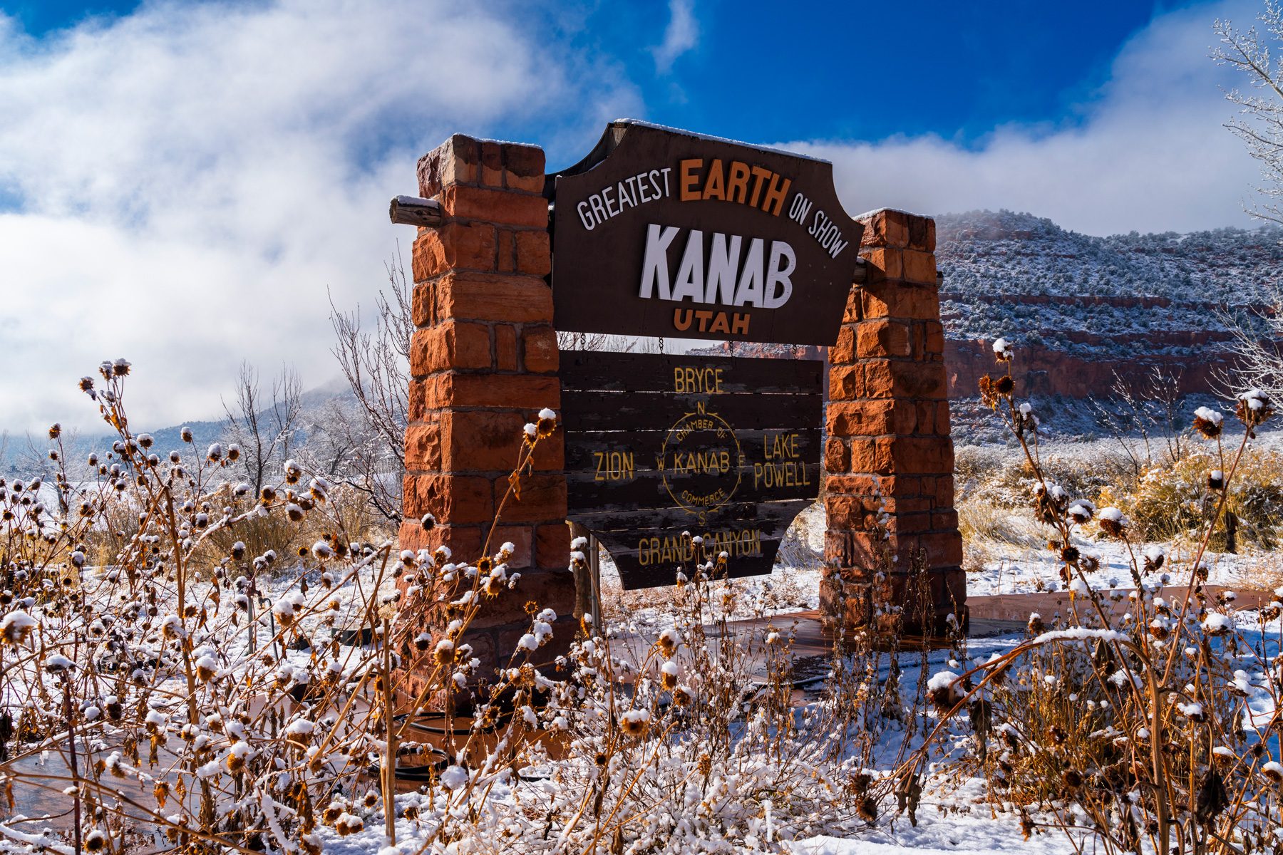 Sign displaying "Kanab, Utah: Greatest Earth on Show," stands amidst snowy landscape and dry plants, beneath a partly cloudy sky.