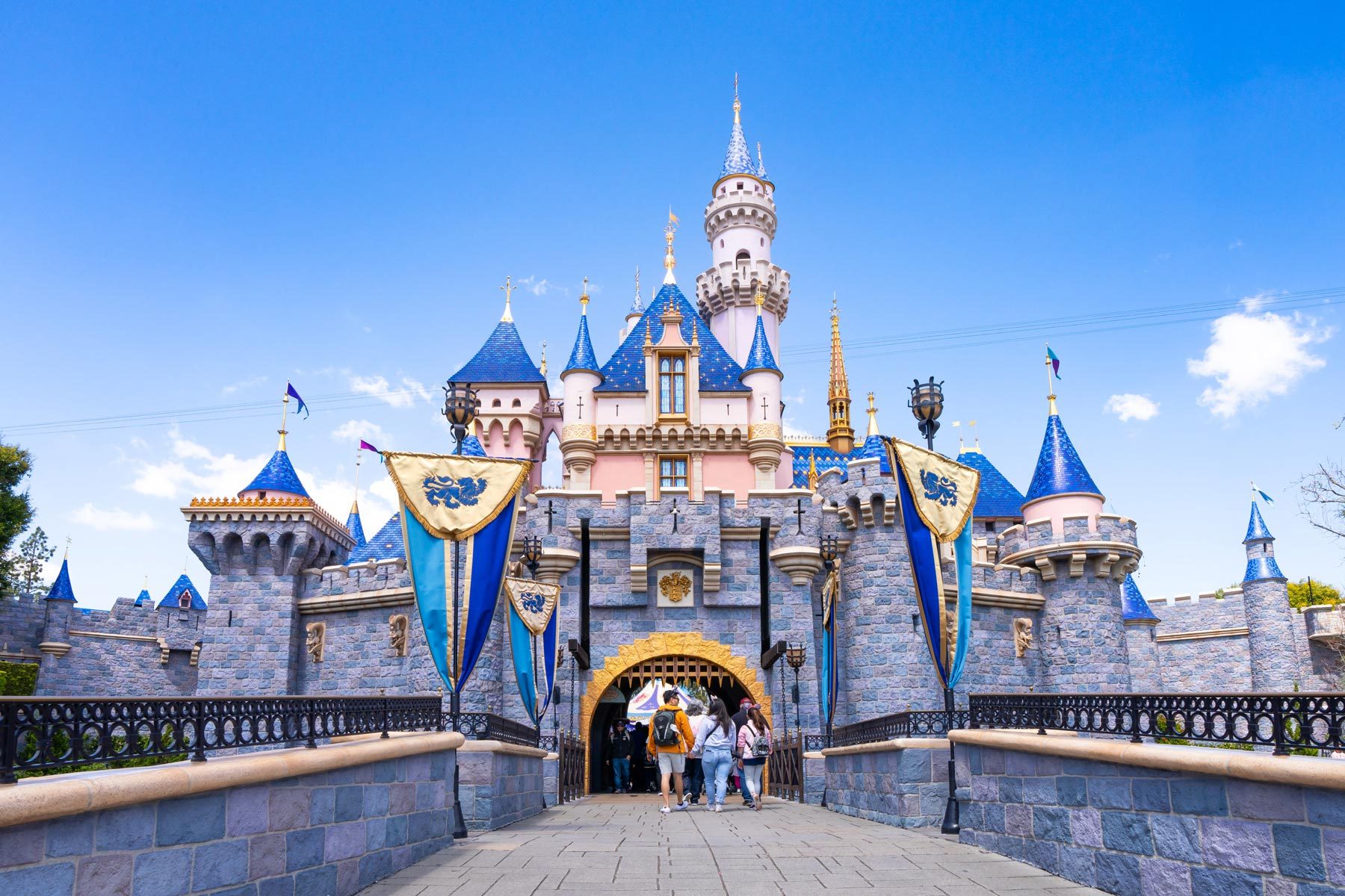 Castle stands tall with visitors entering, surrounded by blue turrets and banners, under a clear blue sky.