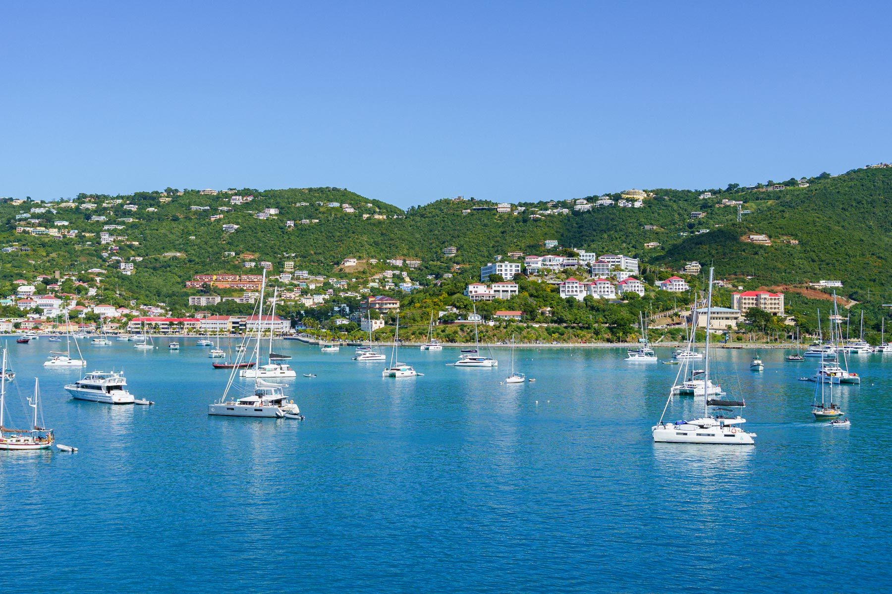 Boats float peacefully on clear blue water, set against lush green hills with scattered buildings beneath a clear blue sky.