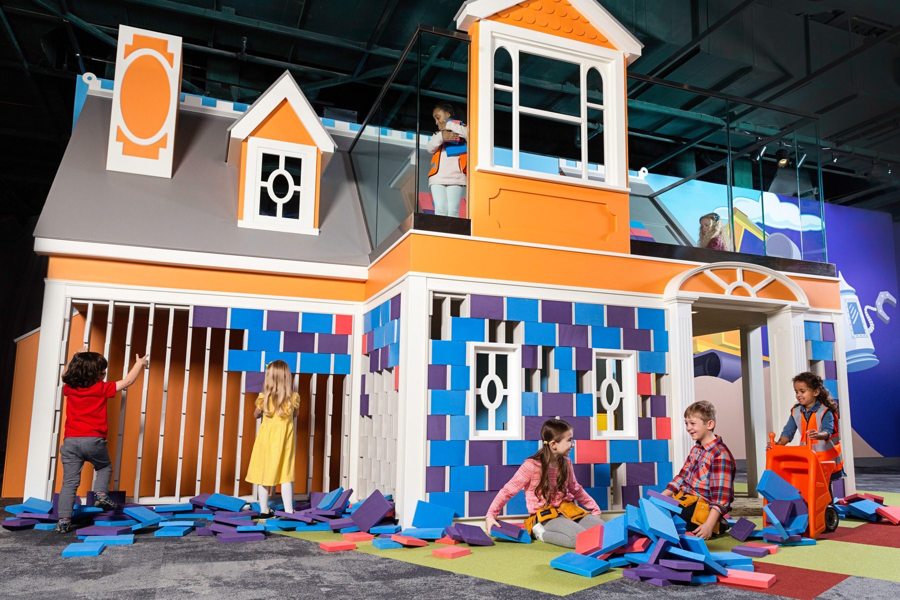 Children play with colorful foam blocks constructing a house structure inside a vibrant, playful indoor environment.