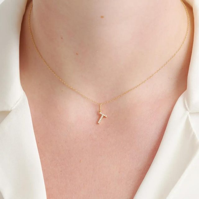 A small gold necklace with a cross pendant rests on a neck, worn with a white shirt in a close-up view.