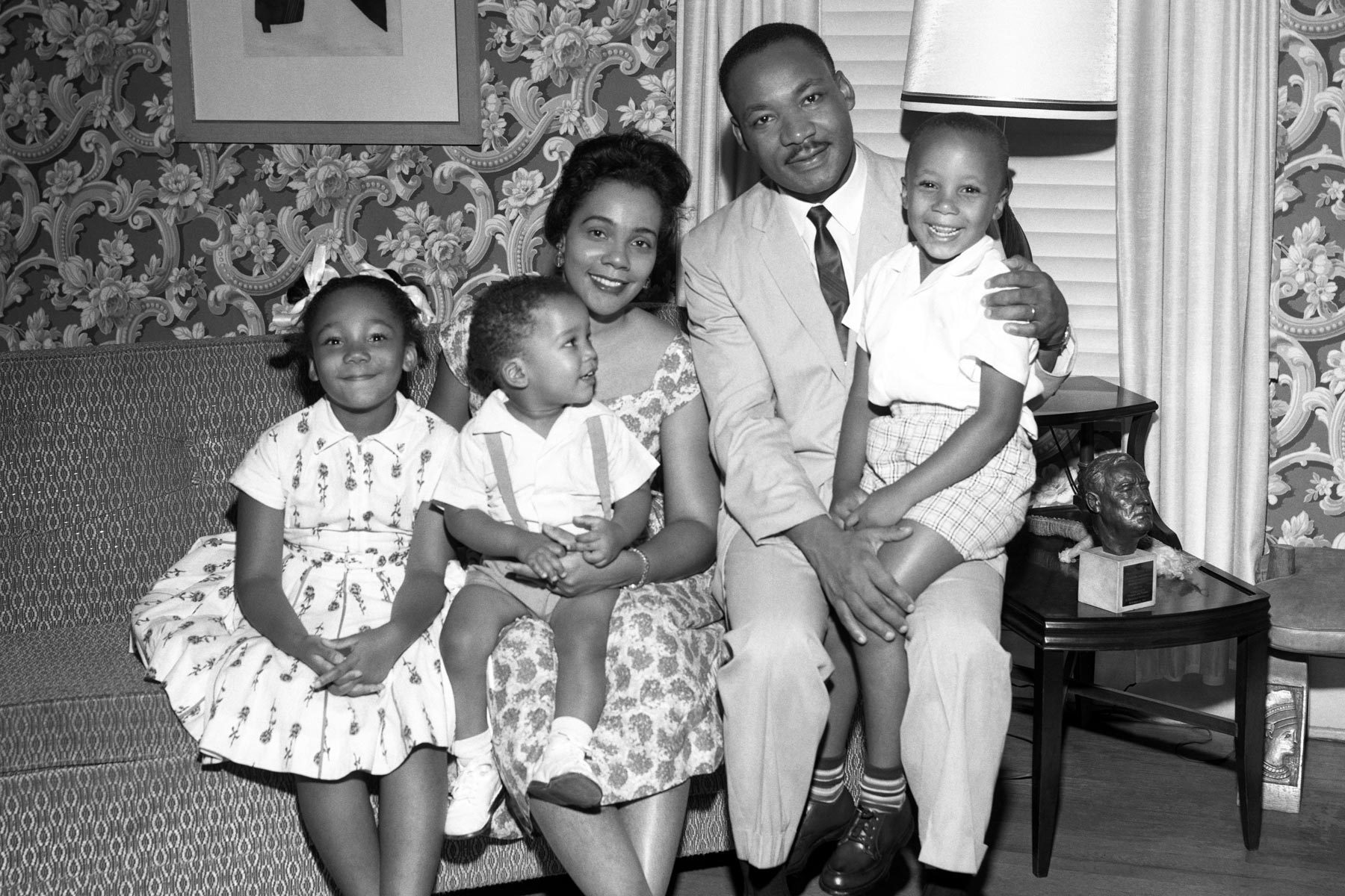 A family sits together on a couch, smiling warmly in a decorated living room with floral wallpaper and a lamp on a side table.