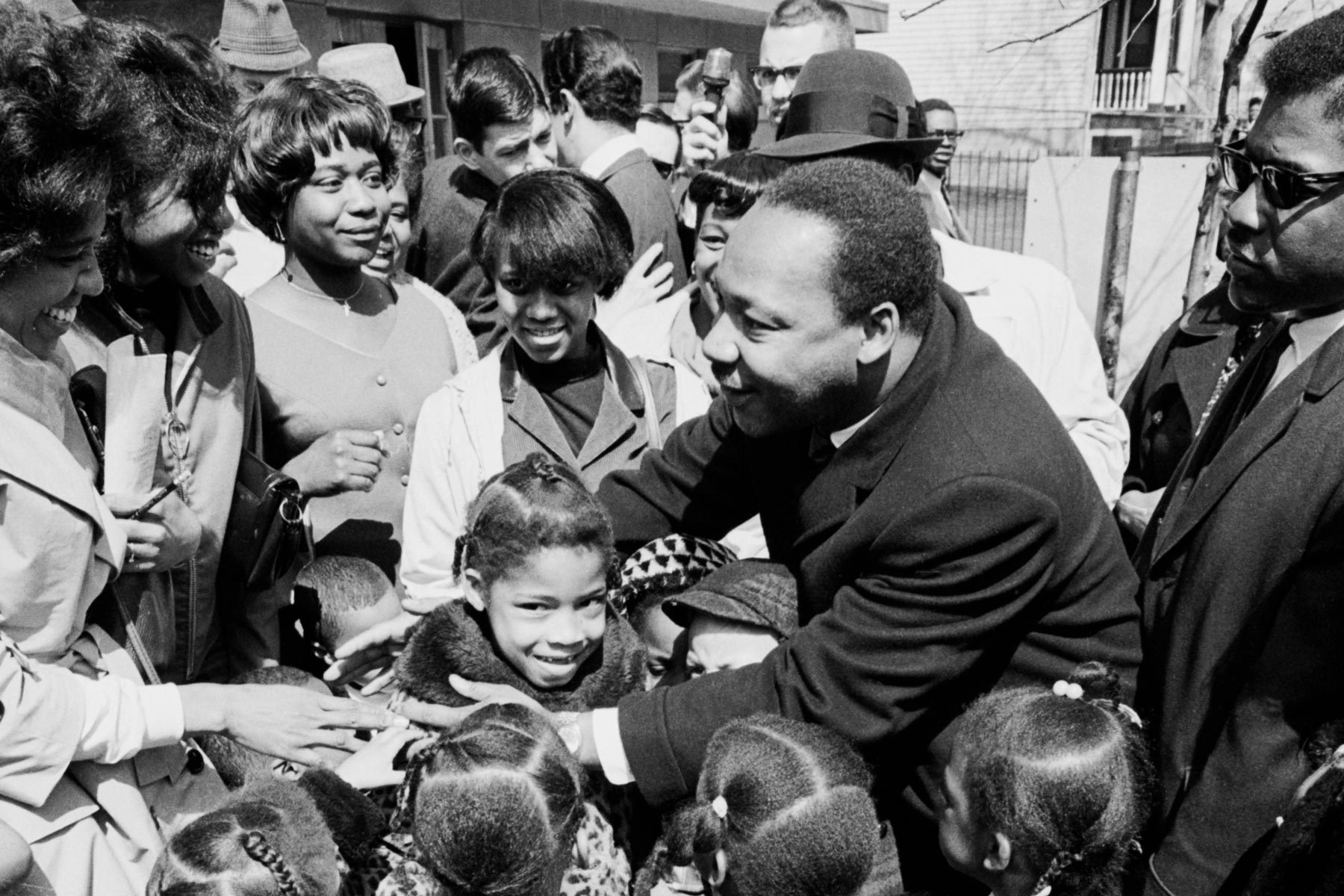 A man smiles and embraces children in a crowded outdoor gathering. Observers are smiling and interacting warmly in a sunny setting.