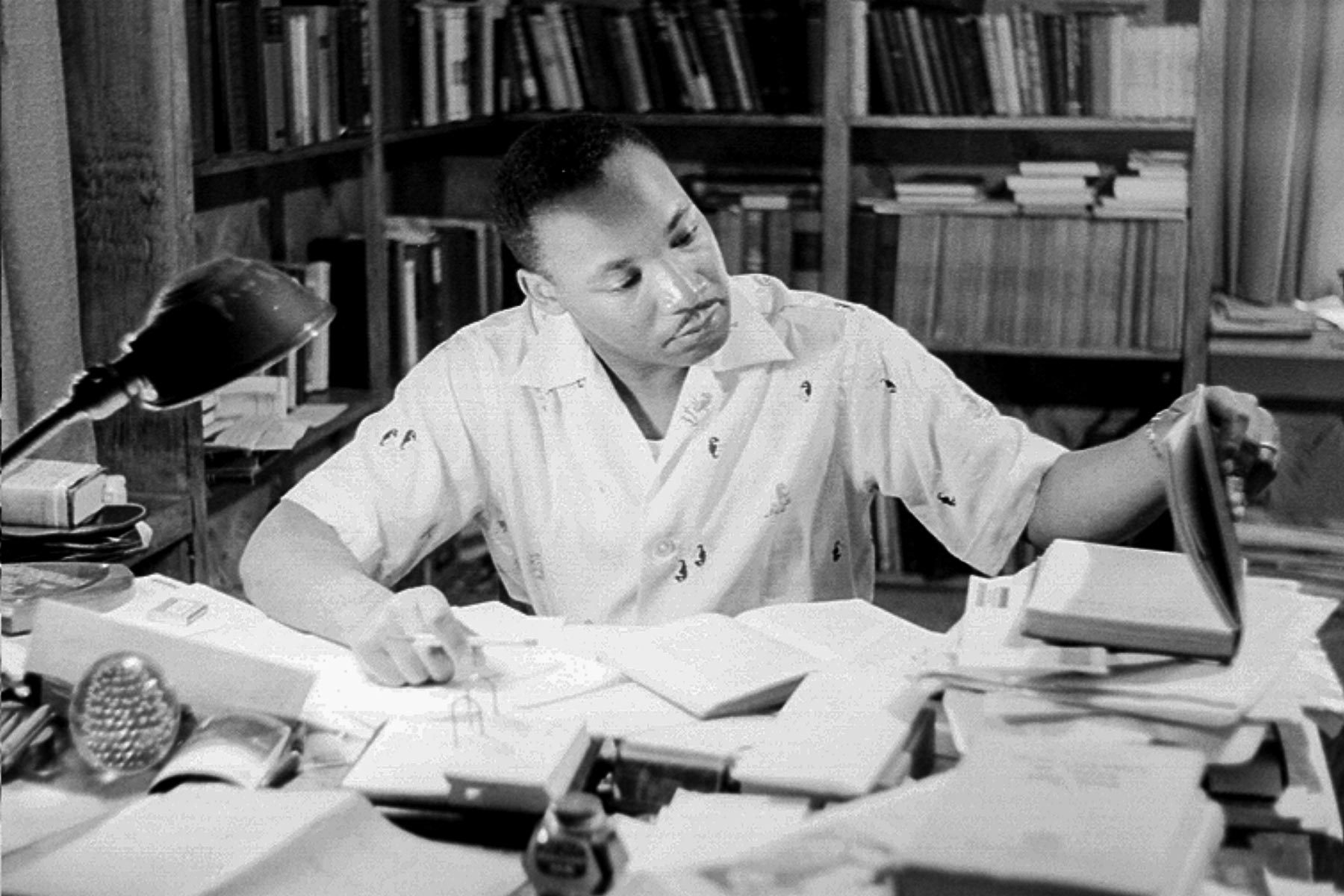 A person reads at a cluttered desk, surrounded by books and papers in a study room with filled bookshelves.