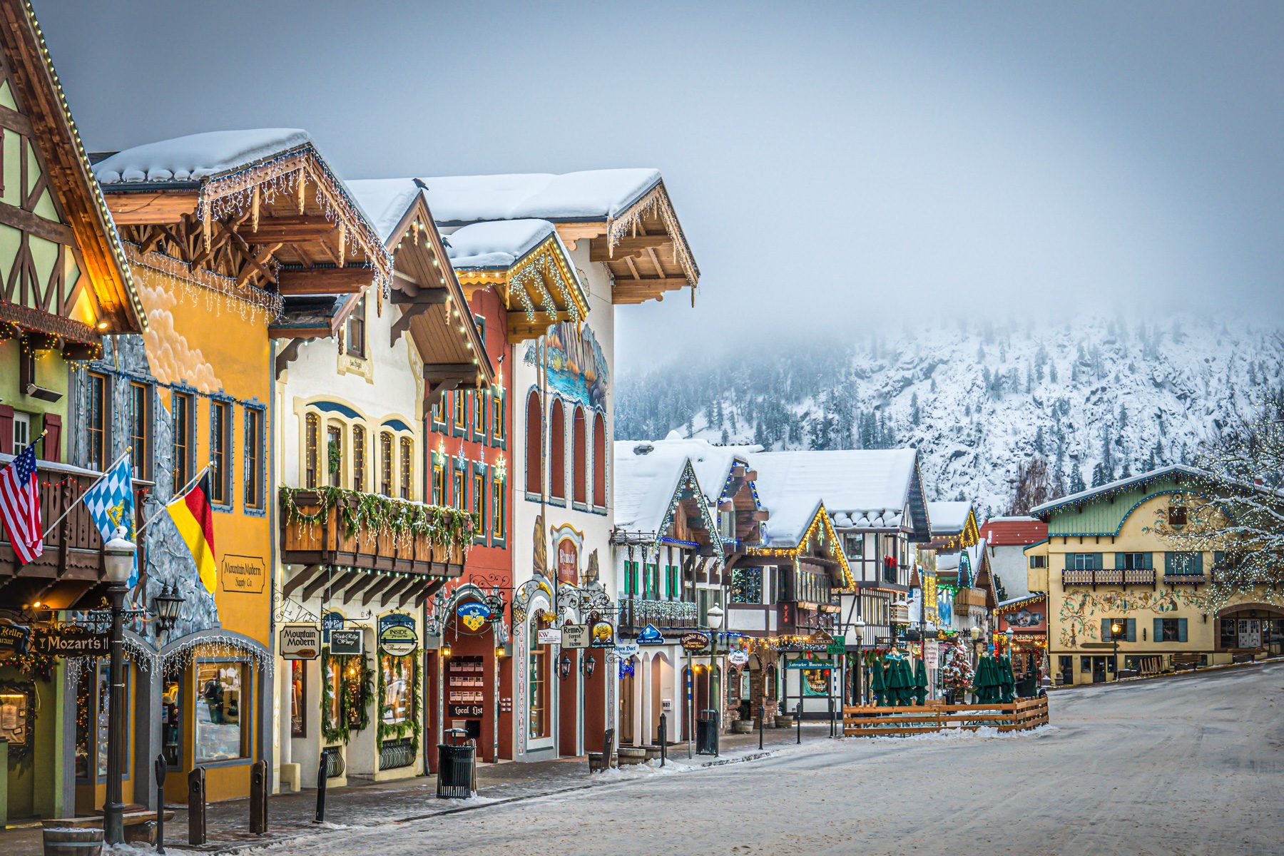 Colorful Bavarian-style buildings stand festively adorned with snow and lights, lining a street in a quaint village, framed by snowy mountains.