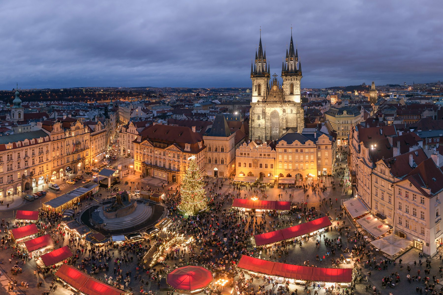Market stalls glow with lights, surrounded by people, in a festive evening square dominated by a grand illuminated church under a cloudy sky.