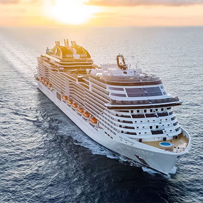 Cruise ship sails on calm ocean at sunset; the sky is orange with sunlight reflecting on water.
