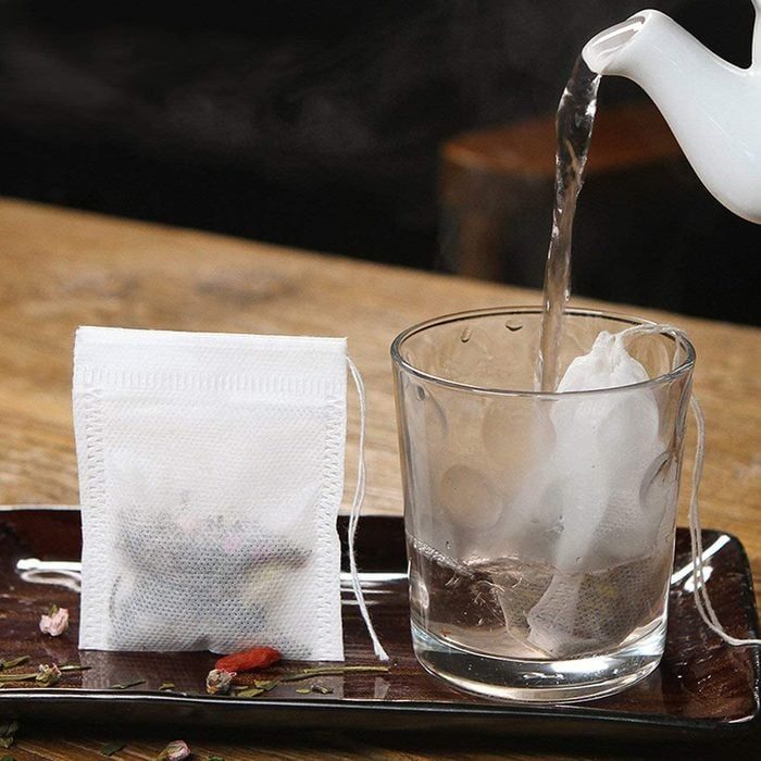 Teapot pours hot water into a glass with a tea bag; another tea bag sits nearby on a wooden tray.