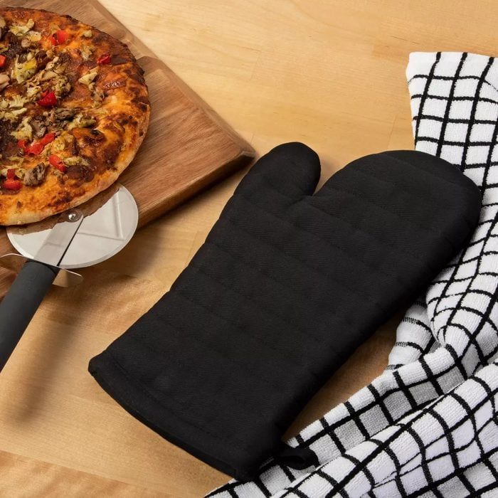 Pizza sits on wooden board beside a pizza cutter, black oven mitt, and checkered cloth on a light wooden table.