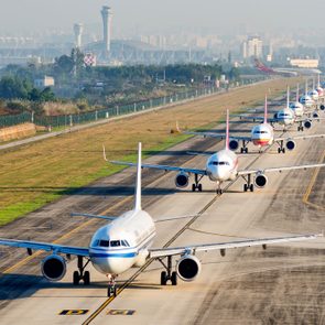 Airplanes lined up neatly on a runway, waiting to take off, surrounded by grassy fields and a distant cityscape.