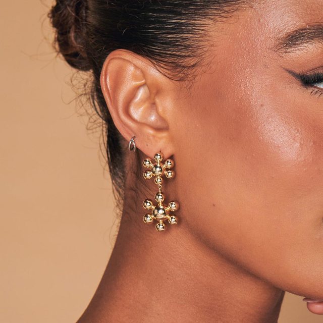 Gold earrings hang, reflecting light, against warm skin and a brown background.
