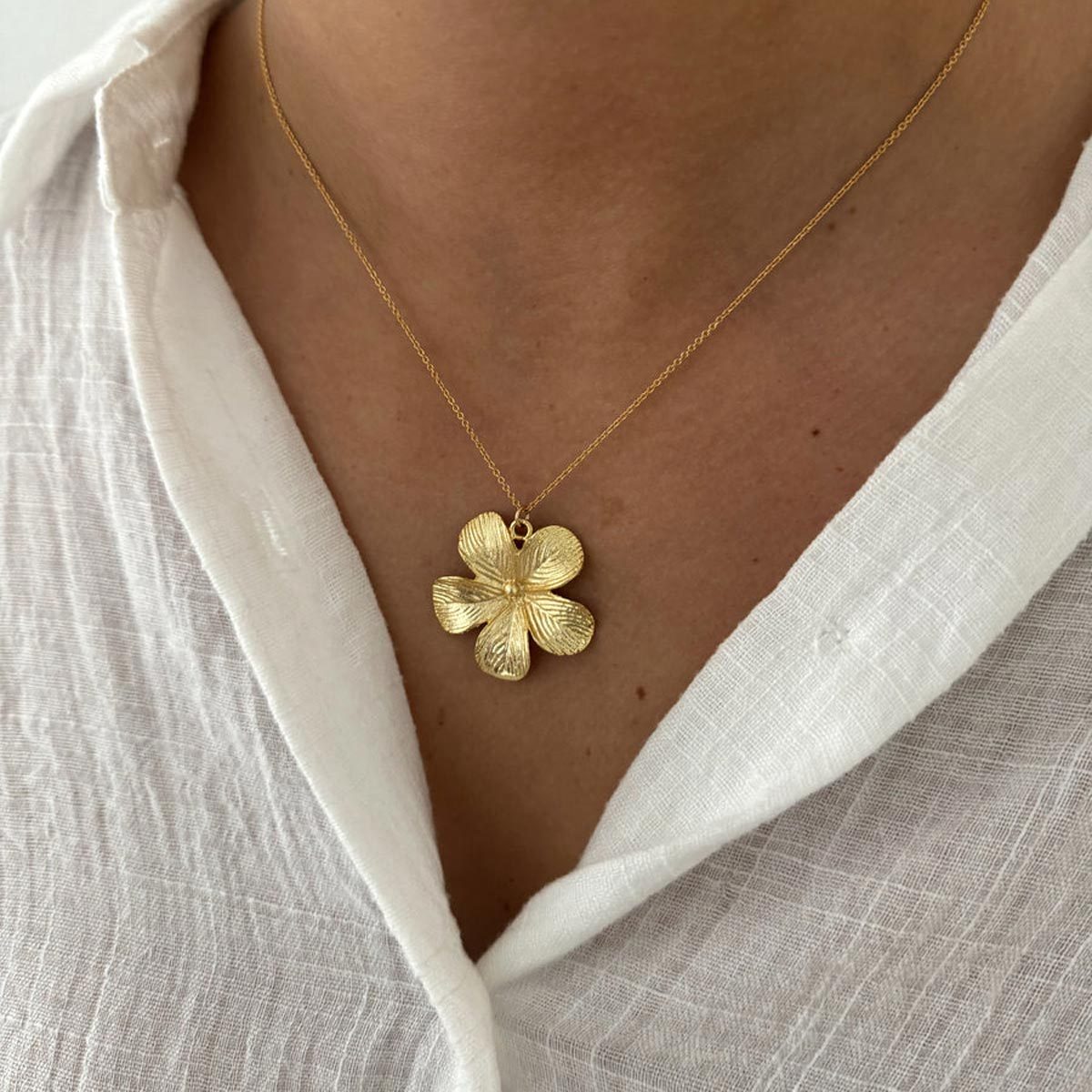 A gold flower pendant hangs on a delicate chain, resting against a person's chest wearing a white textured shirt.
