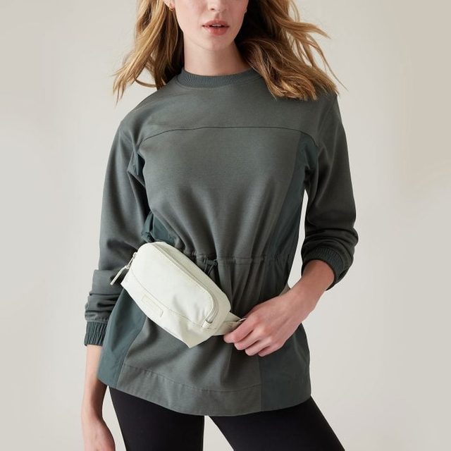 A person stands wearing a green pullover and black pants, holding a white waist bag, against a plain light background.