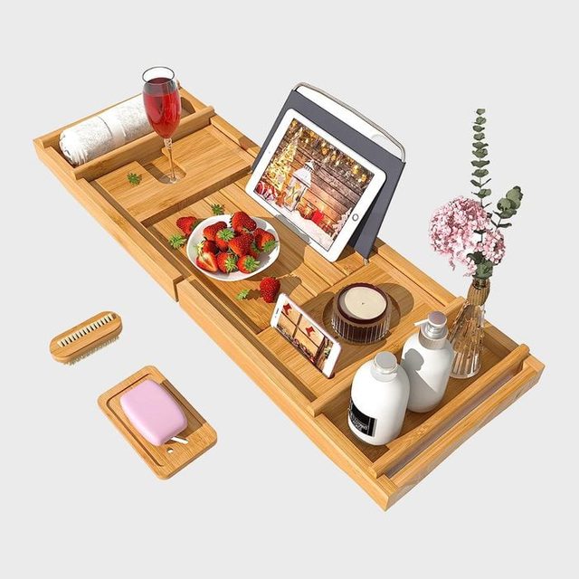 Wooden bath tray holds a tablet, wine, strawberries, phone, candles, soap, and flowers, creating a relaxing bath setting.