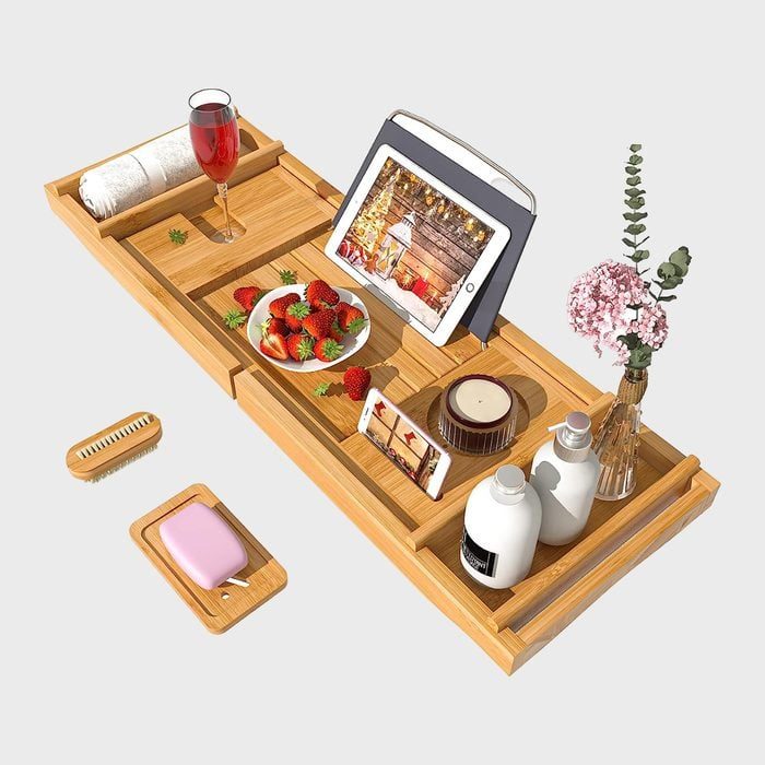 Wooden bath tray holds a tablet, wine, strawberries, phone, candles, soap, and flowers, creating a relaxing bath setting.