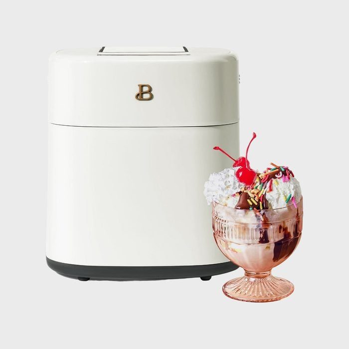 Cream-colored appliance sits beside an ice cream sundae topped with whipped cream, cherries, and sprinkles, on a simple gray background.