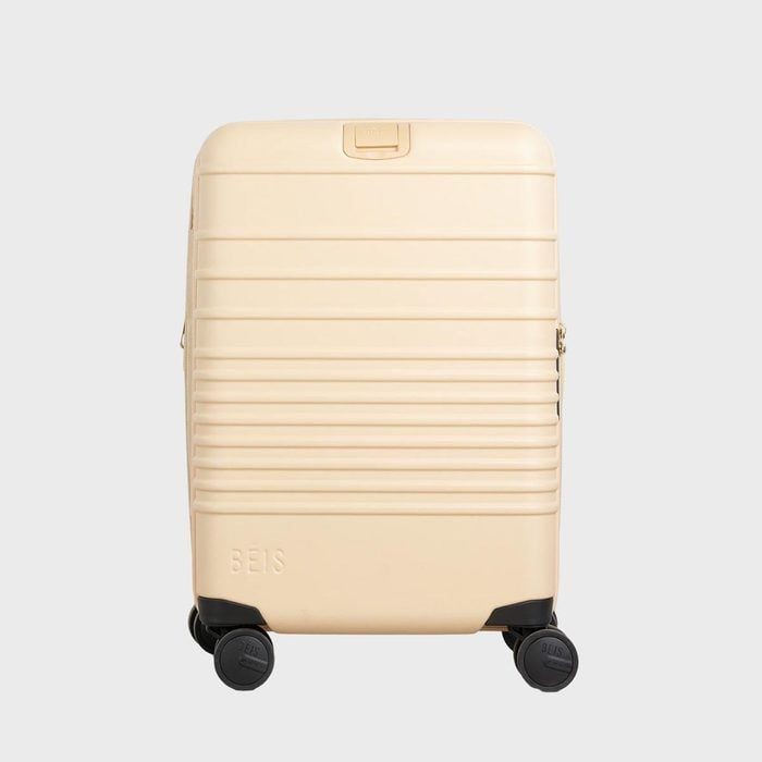 Beige suitcase stands upright, featuring horizontal ridges and four black wheels, against a plain gray background. 