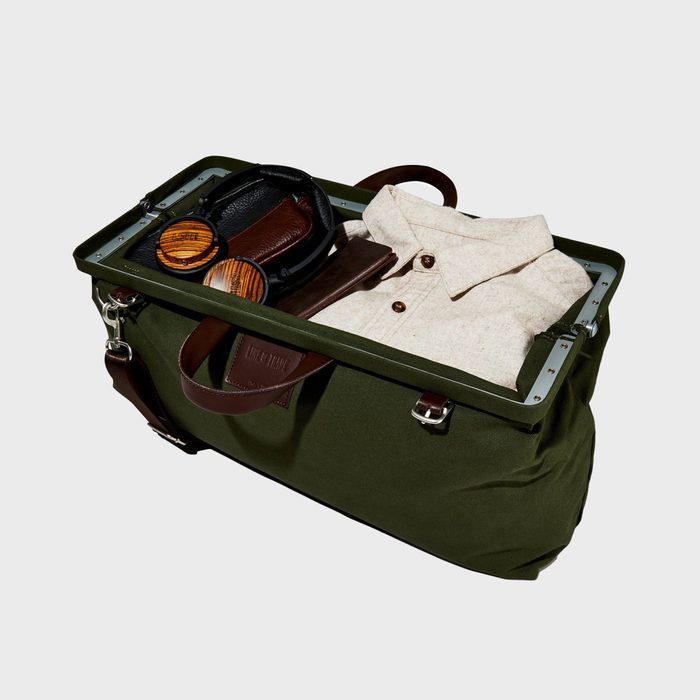 Bag holds folded shirt and headphones; leather wallet and round wooden objects accompany it. Set against a plain, neutral background.