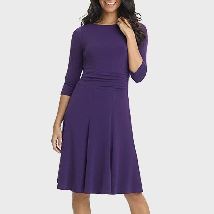 A woman stands smiling, wearing a purple knee-length dress with long sleeves, set against a plain light background.