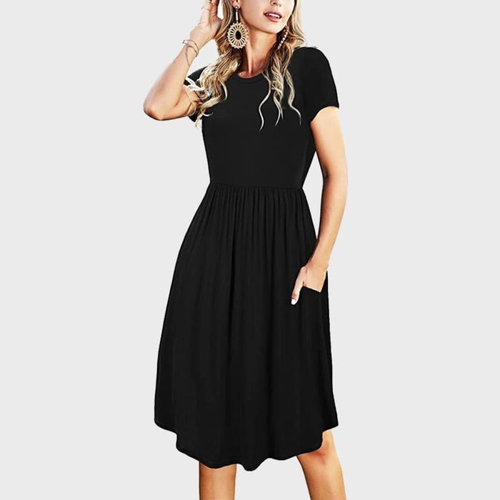 A woman wears a black, short-sleeved dress with pockets, in a minimalistic setting, adjusting her hair with one hand.