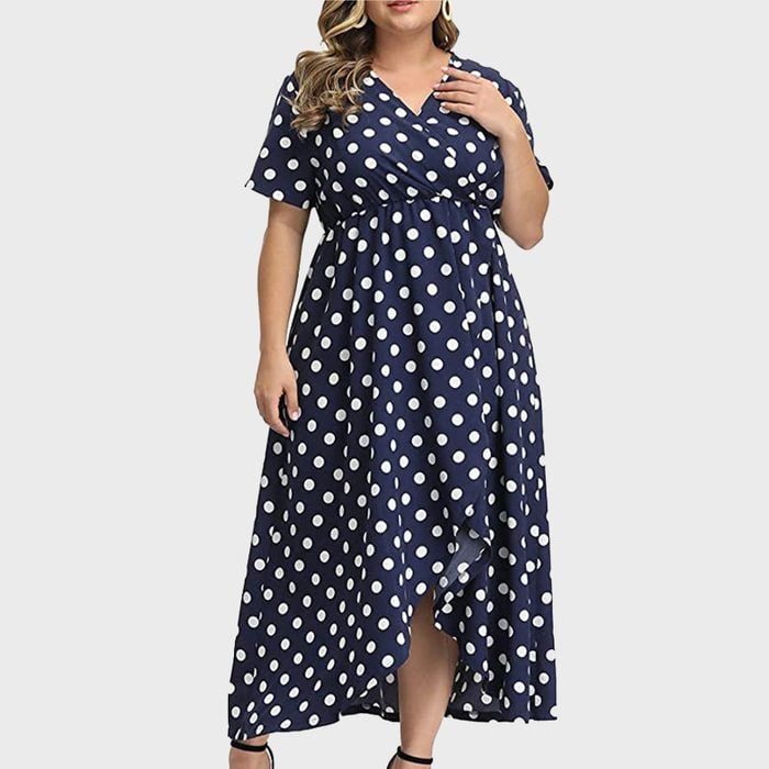 A person wearing a navy-blue polka dot dress stands against a plain light background, hands gently holding the garment.