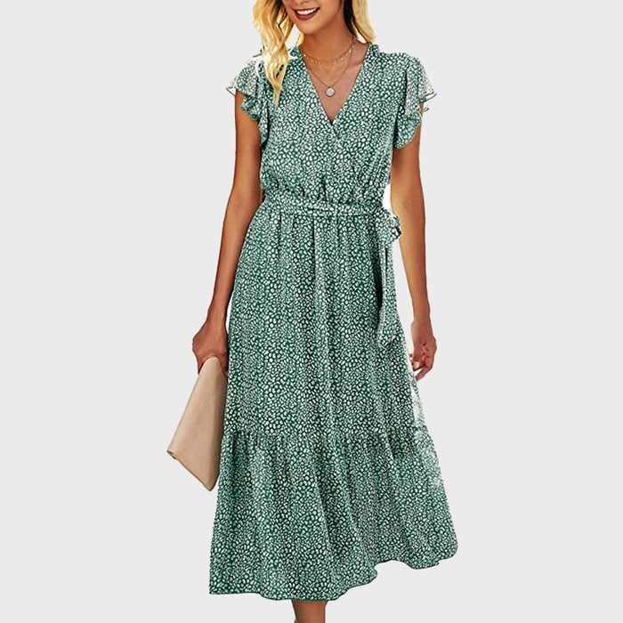 Woman wearing green floral dress, holding a beige clutch, standing against a plain background.