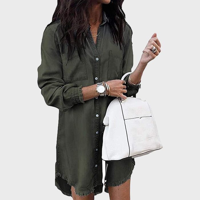 Person stands holding a white handbag, wearing a green buttoned coat-dress with frayed hem, against a plain light gray background.