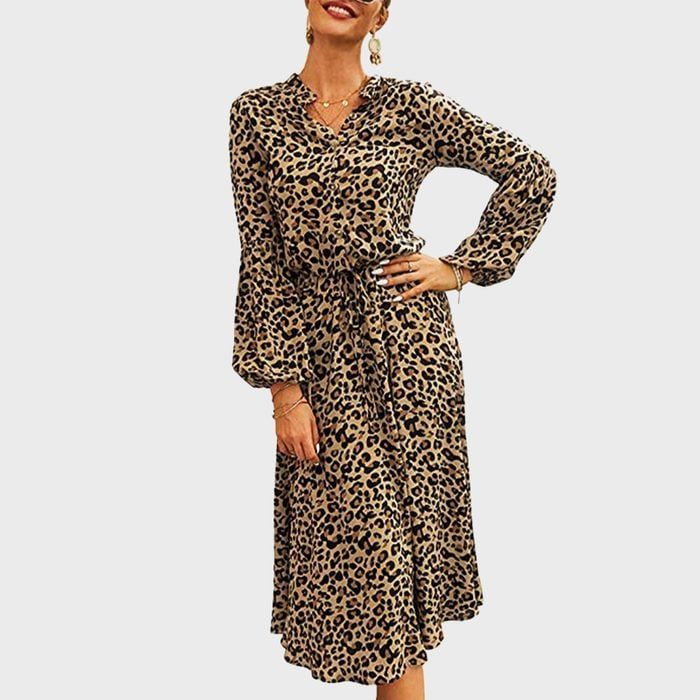Woman wears leopard print dress, smiling with hand on hip; neutral background.