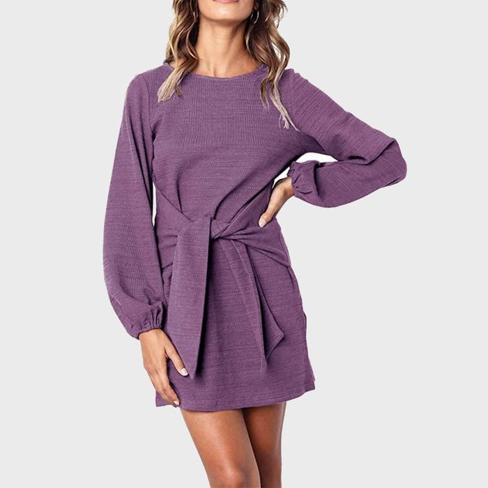 A woman wearing a purple long-sleeve dress with a front tie stands with hand on hip against a plain gray background.