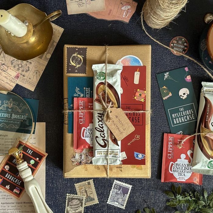 A wrapped book sits with tied chocolate bars and cards, surrounded by vintage stamps, a candle, and twine on a dark fabric.