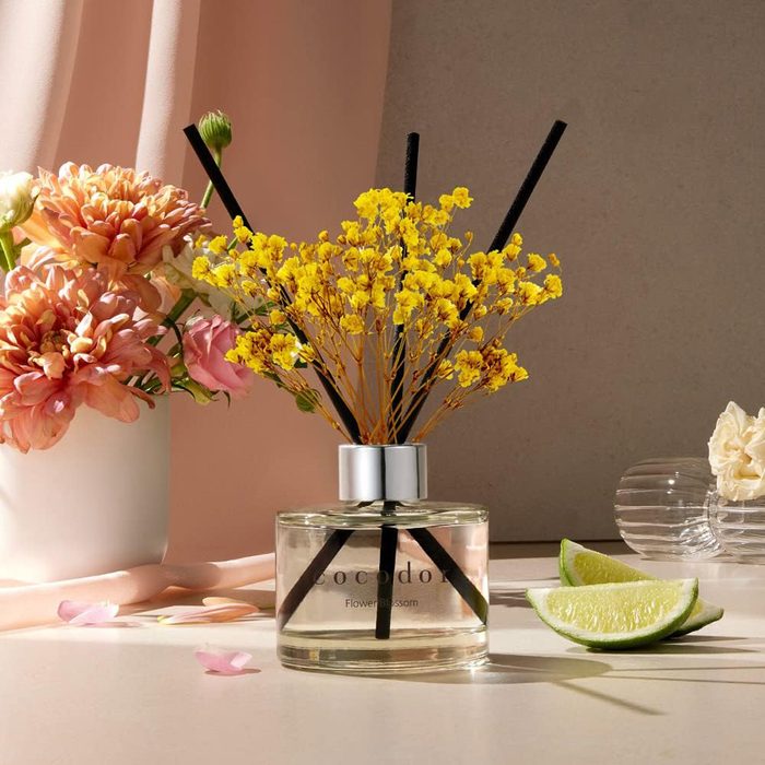 Perfume diffuser displays yellow flowers with black reeds beside pink flowers and lime slices on a sunlit surface.