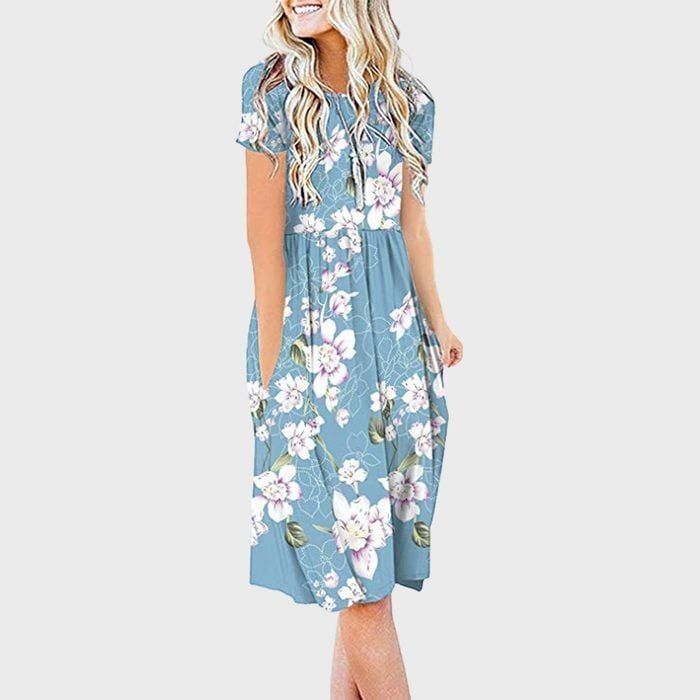 A woman in a blue floral dress stands smiling, with hands in pockets, set against a plain gray background.