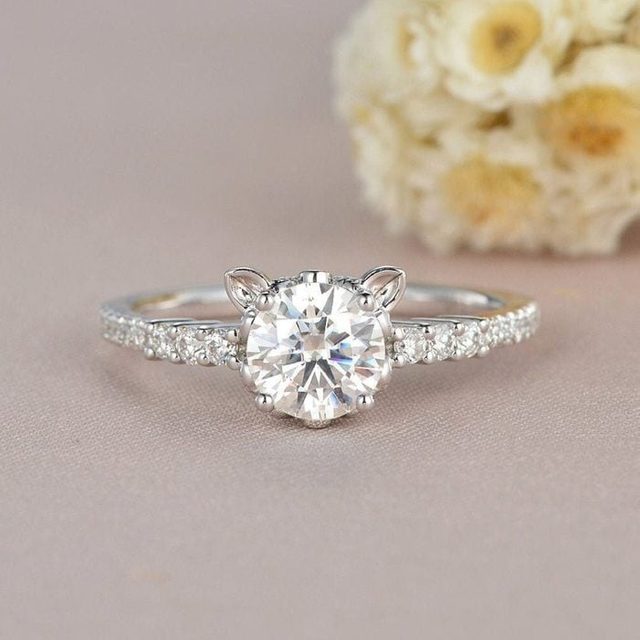 A diamond ring rests on a soft surface, featuring cat-ear designs and a sparkling band; blurred flowers are in the background.