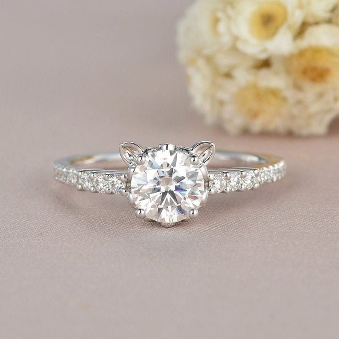 A diamond ring rests on a soft surface, featuring cat-ear designs and a sparkling band; blurred flowers are in the background.