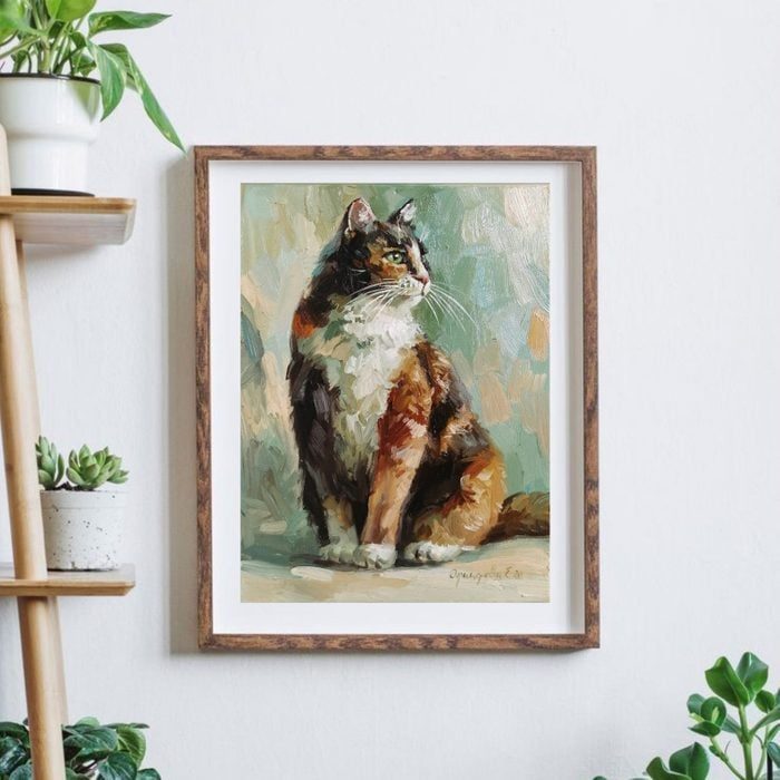 Framed painting depicts a sitting calico cat in an abstract style, hung on a white wall near a wooden shelf with potted plants.