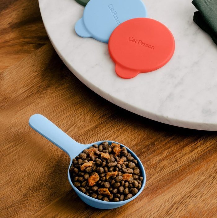 Blue scoop holds cat food on wooden surface; colored lids with 