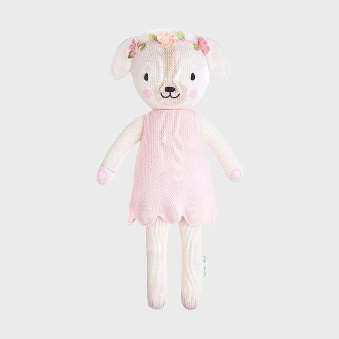 Stuffed teddy bear wears a pink dress and floral headband, standing against a plain gray background.