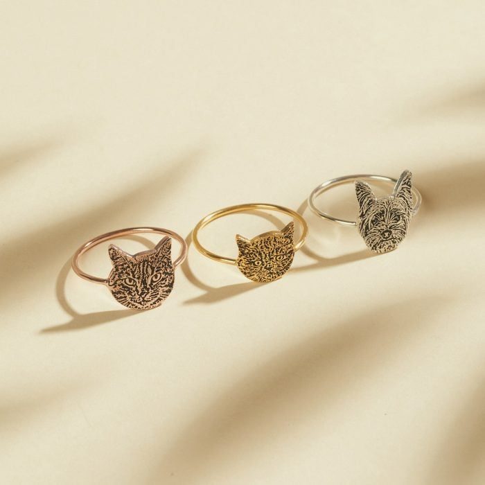 Three cat-faced rings lay side by side on a light beige surface, casting soft shadows in bright, diffused sunlight.