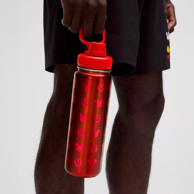 A red water bottle, covered in Mickey Mouse icons, held by a person wearing black shorts, standing in a neutral indoor setting.