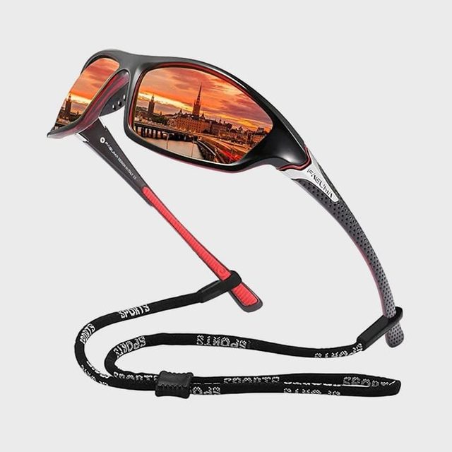 Sunglasses reflect a vibrant cityscape during sunset, featuring buildings and a river. The frame is black with red accents, accompanied by a black sports strap.