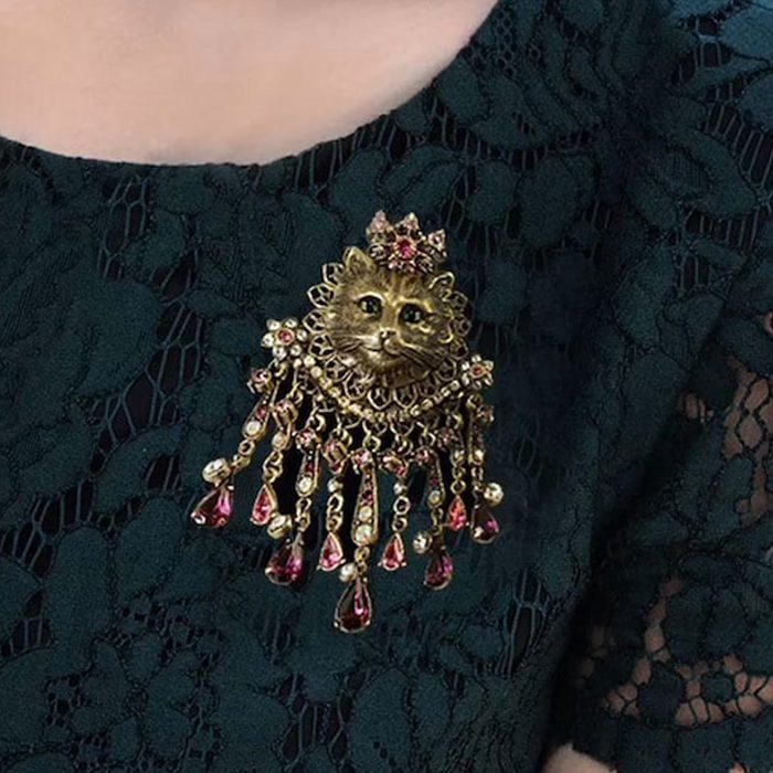 Brooch depicting a cat face adorned with jewels hangs on a dark green lace garment.